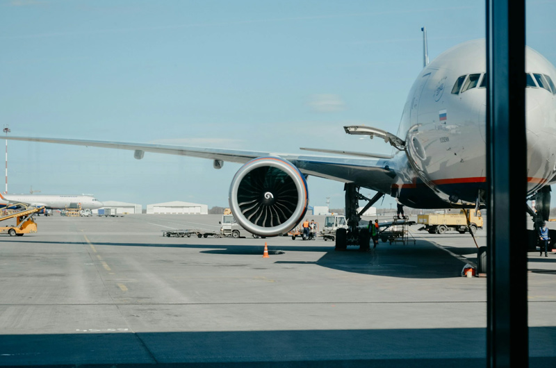A large commercial airplane is parked at an airport gate, with its engine and part of the fuselage visible. Ground service vehicles and airport buildings can be seen in the background on a clear day.