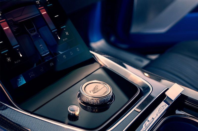 Close-up of a car's center console featuring a large touchscreen display with climate controls and a metallic rotary gear shifter engraved with a logo