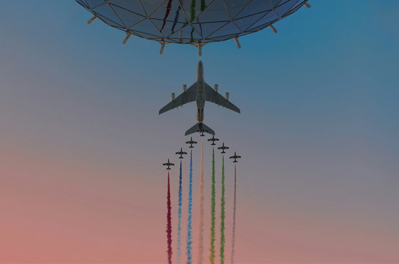 A large airplane and several smaller jets fly in formation, releasing red, blue, green, and white smoke trails against a sunset sky, with a futuristic dome structure above them.