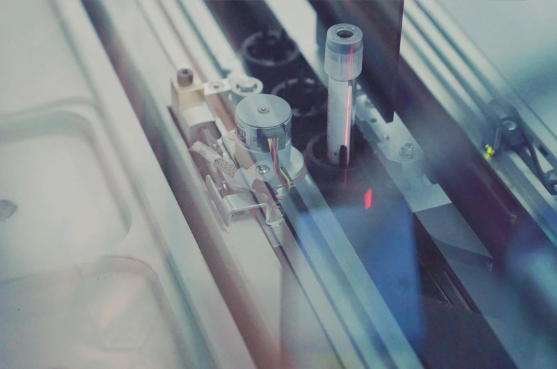 Close-up view of laboratory equipment featuring metal parts and tubes, possibly used for automated chemical analysis or sample processing, under a clear protective cover.