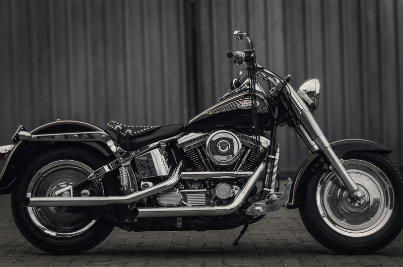 A classic black and chrome motorcycle parked on a paved surface, positioned in profile view, with a metal fence or wall in the background.