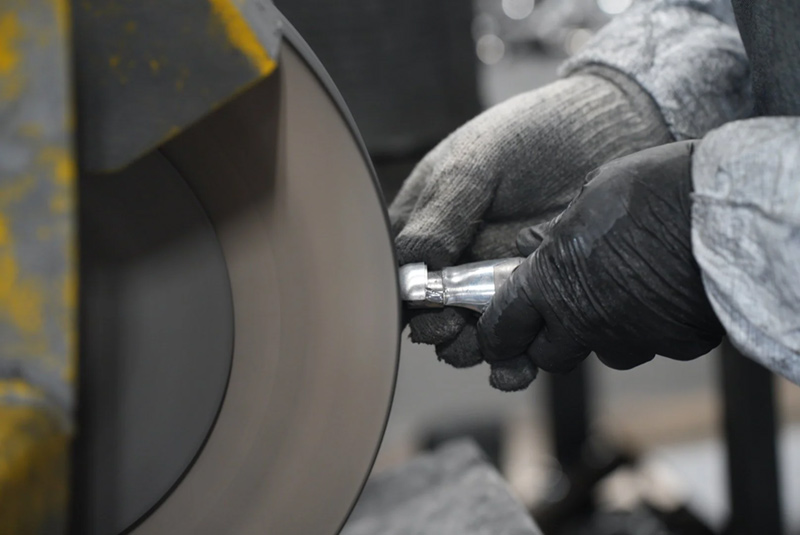 A person wearing gloves holds a small metal object against a large, rotating grinding wheel, polishing the object in an industrial setting.