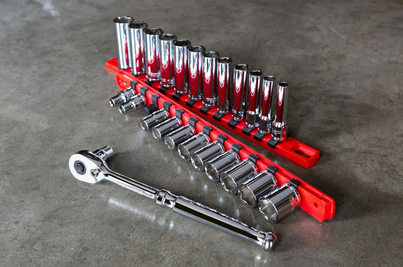 A set of shiny metal socket wrenches on a red holder is arranged on a gray concrete surface, with a ratchet wrench placed in front of the set.